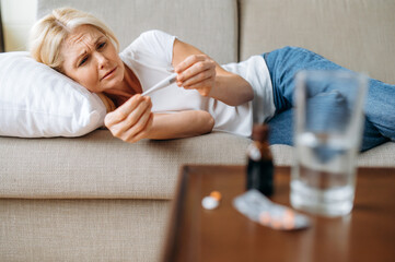 Unhealthy sad mature woman lying on sofa with headache, worried looks at the thermometer, she has a...