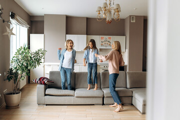 Cheerful caucasian mother, daughter and modern grandmother are having fun at home together, they are dancing, jumping on the sofa, fooling around and laughing