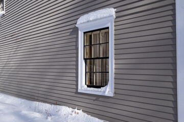 The exterior wall of an old grey building with a small window.  The trim on the building and around the window is white in colour. There's a snowdrift on the top of the window frame and on the ground.
