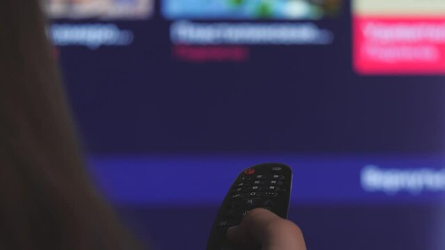 Woman Hand Selects Internet Tv Channels With Remote Control, Close-up. Person Controls TV Using A Modern Remote Control. Girl Watches Smart TV And Uses Black Remote Control. Blurry Tv Scrolls Pages