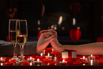 Young couple in love celebrating Valentine’s Day, holding hands lovingly for romantic dinner with red roses, gift, candles and glasses of champagne. Concept about lifestyle, people and celebrations.