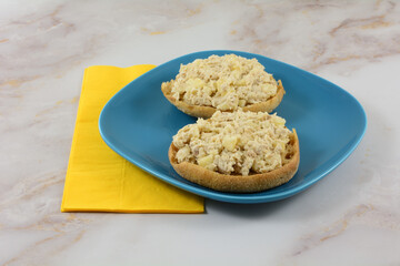 Tuna salad with chopped apple on toasted whole wheat English muffin halves on blue plate with yellow napkin on table