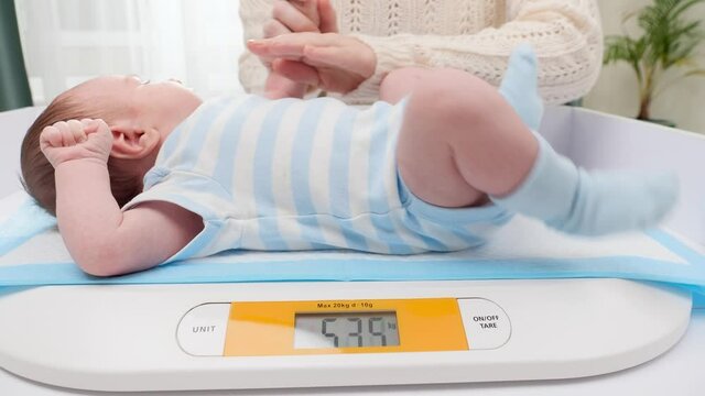 Young caring mother stroking her little newborn baby son while weighing him on digital scales in hospital. Concept of babies and newborn hygiene and healthcare. Caring parents with little children.