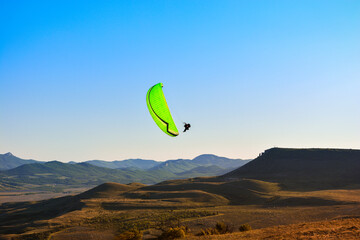 A green paraglider flies over the mountains against the blue sky