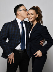 Young couple man and woman in love business professionals office workers in formal suits hug each other and kissing over light wall background. Teamwork, cooperation, business