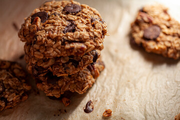 Stacked up fresh made cookies on baking paper. These are made with whole grain rolled oats with dark chocolate chips. There are pieces and crumbles on the paper. Lighting is directional and moody.