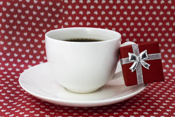 On a red background with hearts there is a white cup of coffee on a saucer. On the saucer is a red gift box. Good morning.Holiday background
