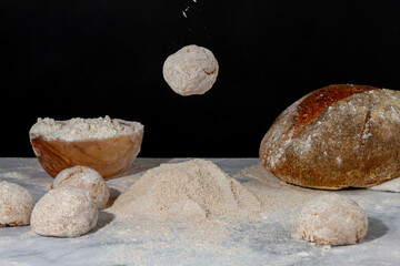 Fototapeta na wymiar A rounded bread dough is falling on to a pile of flour on marble countertop. On the ground there are bread loaves and doughs a bowl of flour and a rustic homemade sourdough bread on muslin cloth.
