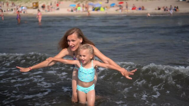 Playful mom and her little daughter facing sea waves. Blurred beach with people