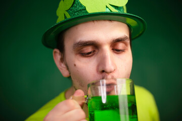 A man in a shamrock hat and green T-shirt drinks a large mug of green ale. Guy celebrates st...