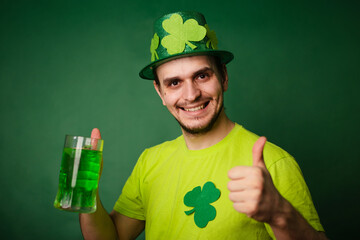 A man in a shamrock hat and green T-shirt drinks a large mug of green ale. Guy celebrates st...