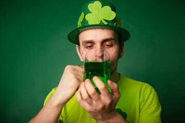 A man in a shamrock hat and green T-shirt drinks a large mug of green ale. Guy celebrates st...