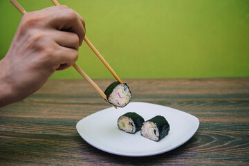 Hand with Chinese chopsticks beats homemade sushi rolls. Rice and fish wrapped in nori seaweed on a white plate.