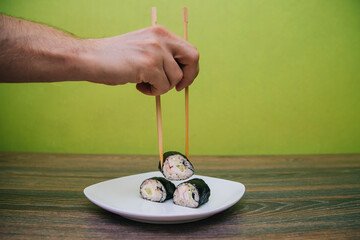 Fototapeta na wymiar Hand with Chinese chopsticks beats homemade sushi rolls. Rice and fish wrapped in nori seaweed on a white plate.