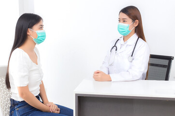 Asian woman doctor is talking with a girl patient at hospital, both of them wear medical face mask to protect microorganism pathogen in respiratory system.