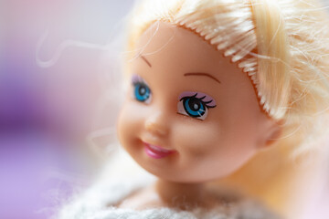 Close up of a small plastic doll with blond hair and blue eyes smiling against a pastel colored...