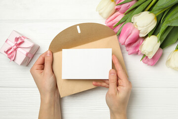 Female hands hold envelope with blank card on white wooden background with tulips and gift box