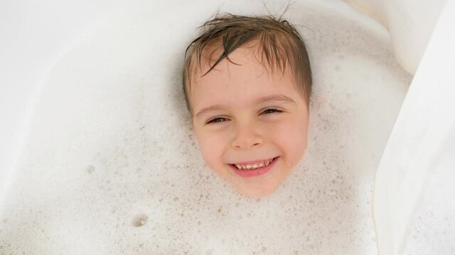 Portrait of cheerful little boy lying in bath with soap foam and looking in camera. Concept of child hygiene and health care at home. Child having fun and playing at home.