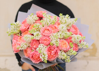 Close up of composition of lilacs and David Austin roses wrapped in pink paper . Delicate bouquet of white lilacs and pink David Austin roses. Man holding fresh beautiful bouquet.