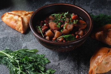 Bean soup with herbs in a clay bowl with fresh rye bread and garlic on a slate table on a dark background.