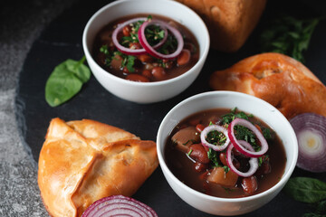 Bean soup with herbs and red onion rings in two small white bowls with fresh rye bread and garlic on a slate table on a dark background.