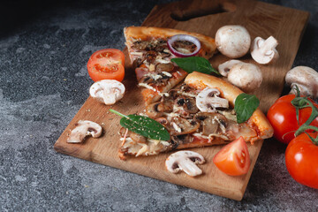 Slices of homemade Italian pizza on a chopping wooden board with mushrooms, ham, tomatoes, cheese, onions and herbs lie on a dark stone slate table, dark background .