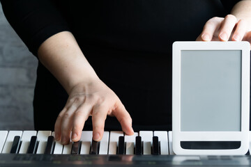 Mock up for online courses play the piano. One hand plays on the keyboard, the other holds the tablet, the screen turned forward. Close-up, selective focus, copy space