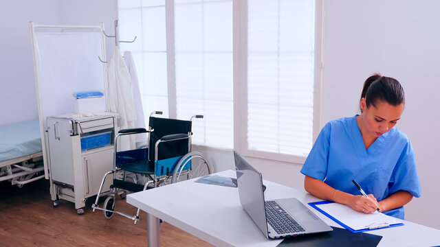 Woman therapist sitting at workplace looking at laptop reading news and taking notes on clipboard. Medical physician in medicine uniform writing list of consulted, diagnosed patients, making research.