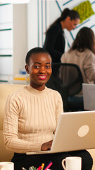 Portrait of african woman typing on laptop looking at camera smiling while diverse team working in background. Multiethnic coworkers talking about startup financial company in modern business office