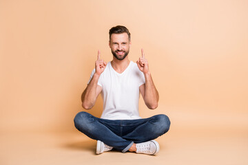 Portrait of nice cheery guy sitting on floor in lotus pose pointing forefinger up copy space ad isolated over beige color background