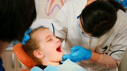 Close up of little patient lying on chair with open mouth during dental examination. Stomatologist holding sterilized tools talking with girl before stomatological intervation. Doctor wearing mask