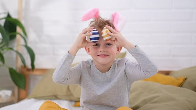 Portrait Easter kid. Boy in rabbit bunny ears on head having fun looking into the camera making crazy funny face with colored Easter eggs on his eyes at home. Cheerful smiling child