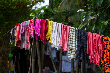 Colorful clothes hanging on rope in green garden. Multicolored textile clothing in asian village. Rustic lifestyle hygiene in countryside. Social issues in South Asia. Big family washing day