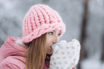 Happy cheerful caucasian girl drinks tea in winter snowy park. Beautiful happy young woman posing outdoors in knitted hat