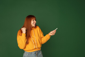 Winner, smiling. Caucasian woman's portrait isolated on green studio background with copyspace. Beautiful female model with phone. Concept of human emotions, facial expression, sales, ad, fashion.