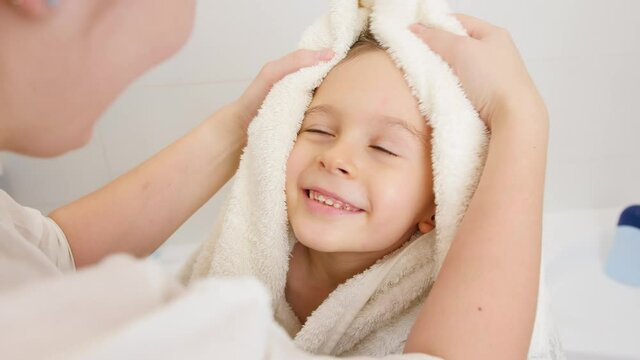 Portrait of cute smiling boy with wet hair wiping and drying with soft towel after having bath and looking on caring mother. Concept of child hygiene and health care at home.