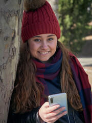 Vertical shot of a young female in winter clothes leaning on a tree and using her smartphone