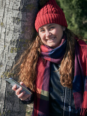 Vertical shot of a young female in winter clothes leaning on a tree and using her smartphone