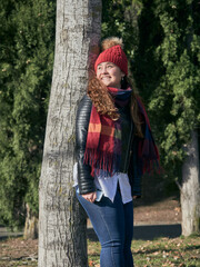 Vertical shot of a happy young female in winter clothes leaning on a tree in the park