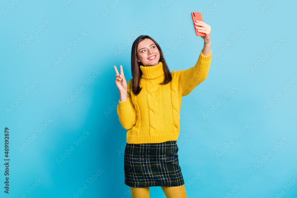 Poster portrait of pretty cheerful girl blogger taking selfie posing showing v-sign isolated over bright bl