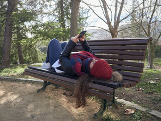 Young caucasian female using her smartphone while lying on a wooden bench in the park