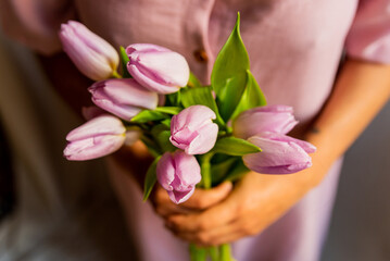 Obraz na płótnie Canvas closeup of lilac tulips in young woman hands with natural soft light and simple pink dress. concept of spring time and femininity. authentic people, natural looking portrait and lifestyle