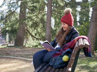 Young caucasian female reading book while sitting on a wooden bench in the park