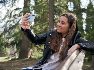 Young caucasian female taking a selfie while sitting on a wooden bench in the park