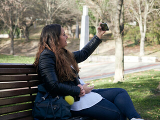 Young female taking a selfie with her smartphone while sitting on a wooden bench in the park
