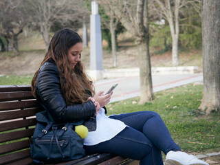 A young caucasian female using her smartphone while sitting on a wooden bench in the park