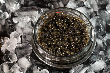 The black caviar of the sturgeon fish in a glass jar is laid on the crushed ice. Top view, copyspace - obrazy, fototapety, plakaty