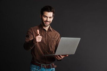 Joyful handsome guy using laptop and pointing finger at camera