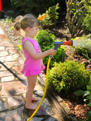 Beautiful little toddler girl watering garden flowers with water hose on summer day. Happy child...