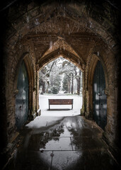 Winter churchyard scene of grave yard stones single bench alone in falling snow. Church archway reflected beautiful wooden doors and pathway peaceful blanket of snowfall in local Victorian cemetery